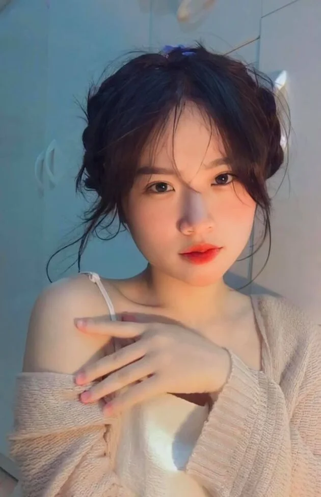 Ảnh gái xinh châu Á tóc búi hai bên, son đỏ, áo len be trễ vai chụp selfie trong phòng